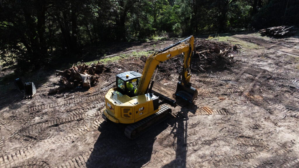 A yellow Cat 310 excavator on a dirt clearing with piles of wood debris and surrounding trees